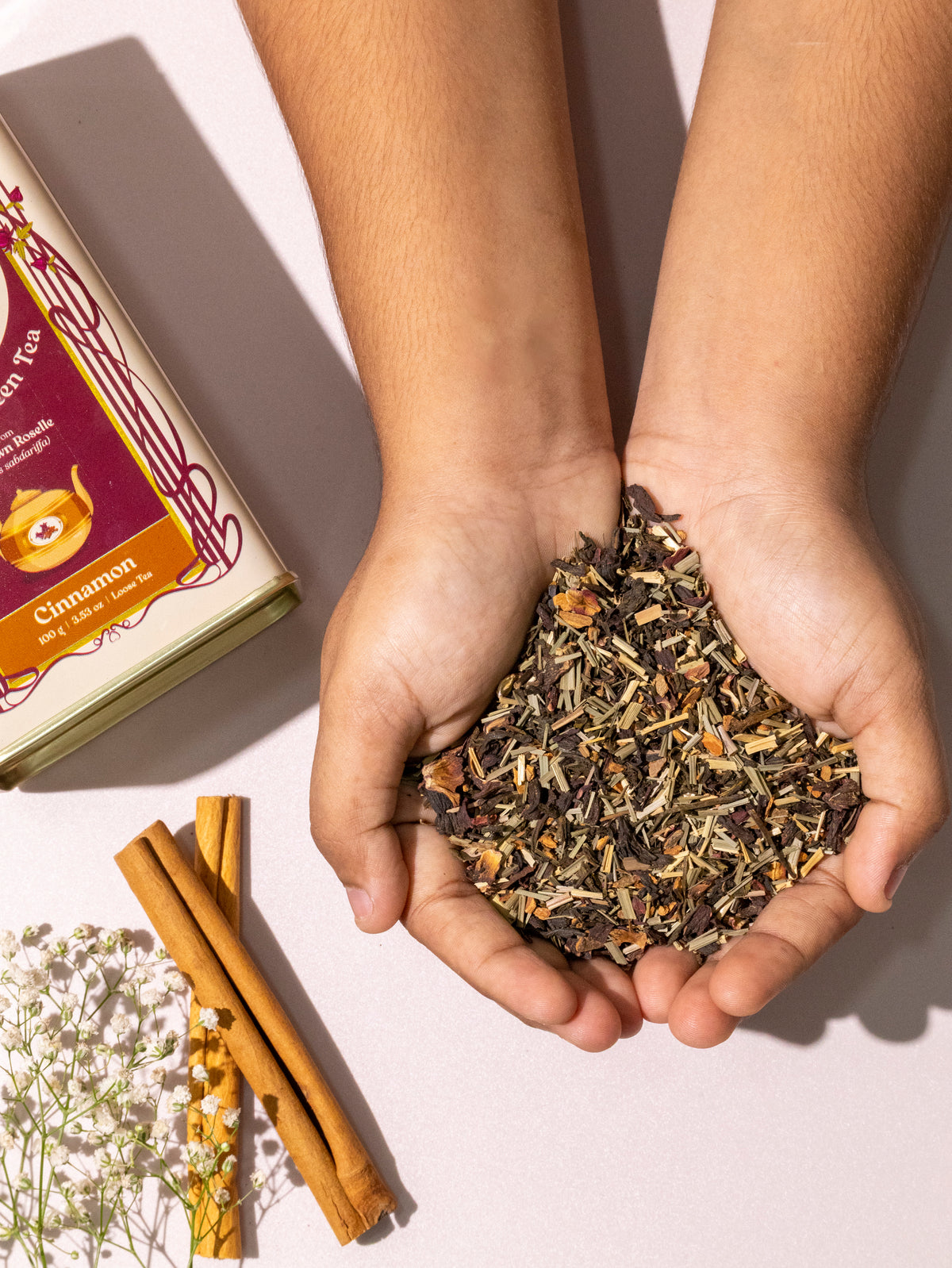 Two hands holding loose roselle green tea leaves beside cinnamon bark